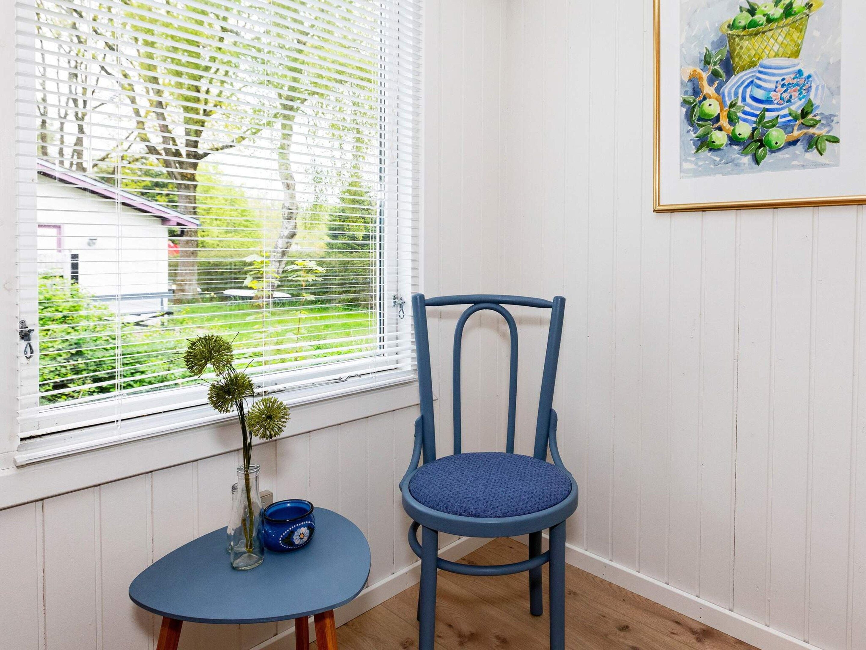 Una habitación con ventana, una silla, una mesita y un cuadro en la pared.