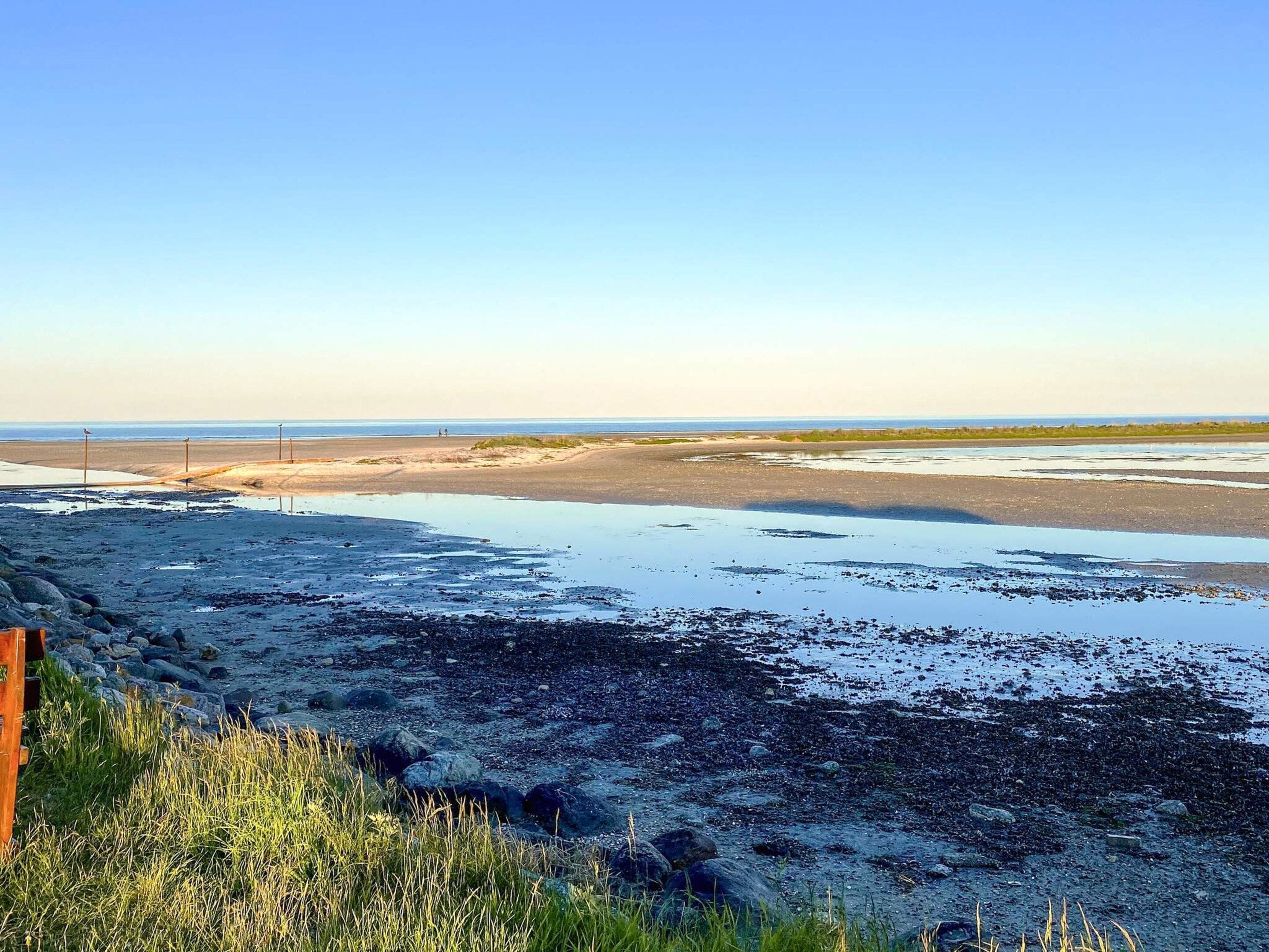 A coastal landscape with a sandy beach, rocky foreground, and a clear sky.