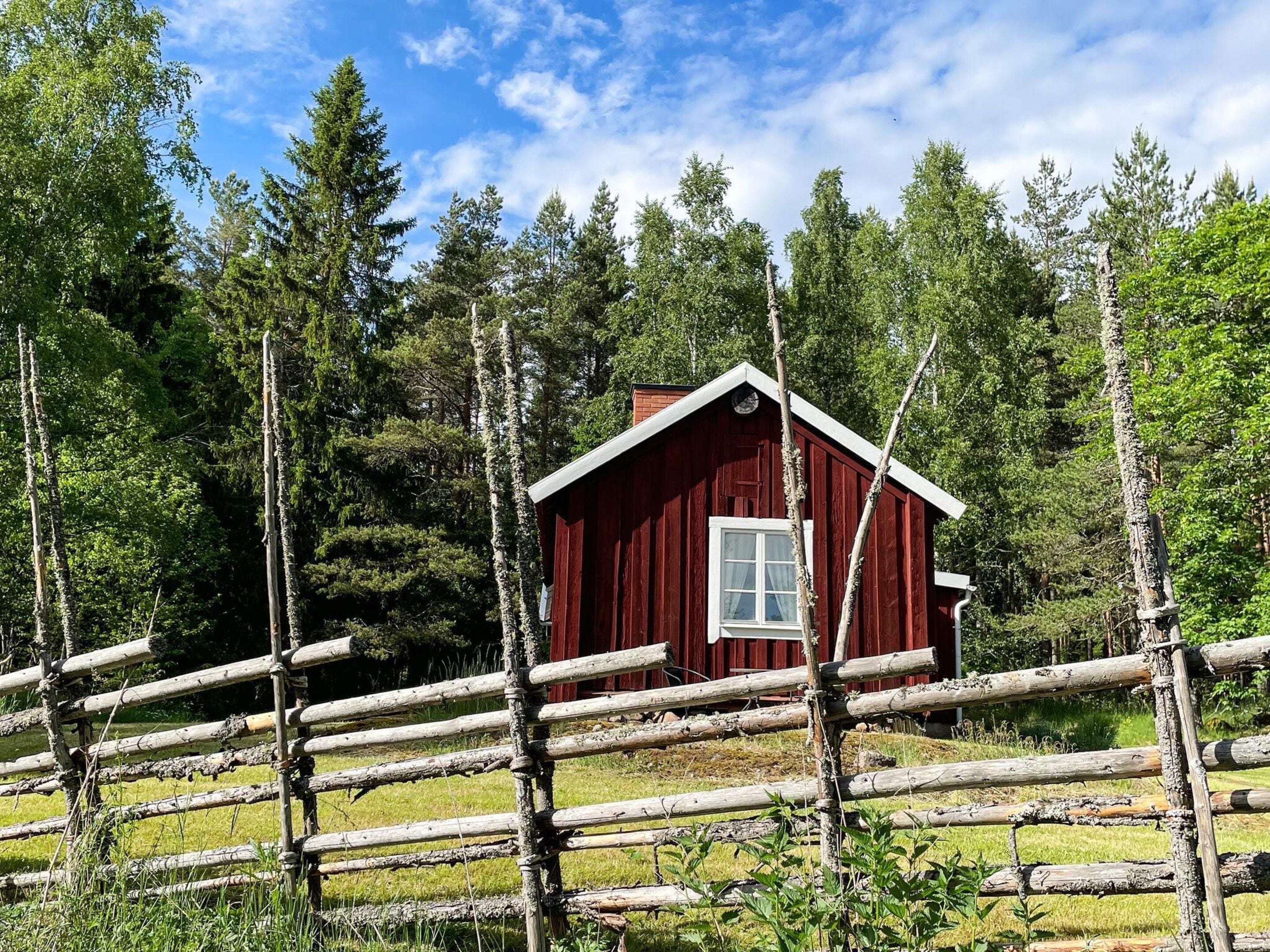 Eine rote Holzhütte mit einem weißen Fenster, umgeben von einem Holzzaun und hohen Bäumen.