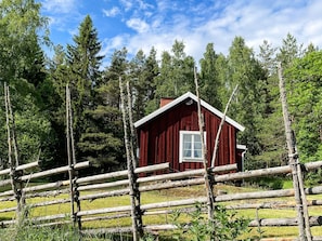 A red wooden cabin with a white window, surrounded by a wooden fence and tall trees. - 3 Person Holiday Home in Eskilstuna (Eskilstuna)