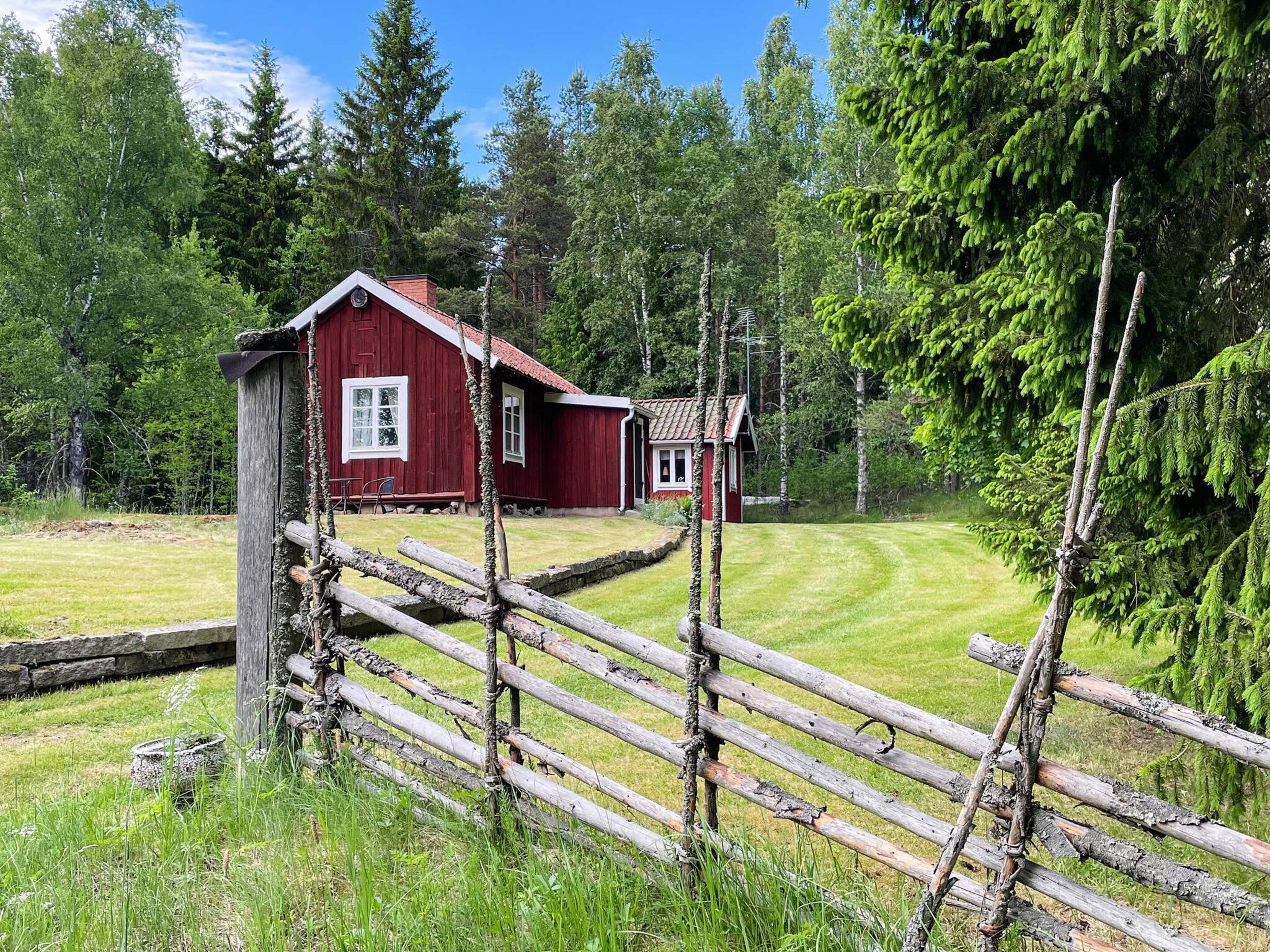 A red wooden cabin with a small attached structure, surrounded by a wooden fence and lush greenery.
