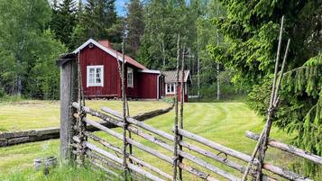 A red wooden cabin with a small attached structure, surrounded by a wooden fence and lush greenery.