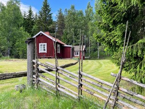 A red wooden cabin with a small attached structure, surrounded by a wooden fence and lush greenery. - 3 Person Holiday Home in Eskilstuna (Eskilstuna)