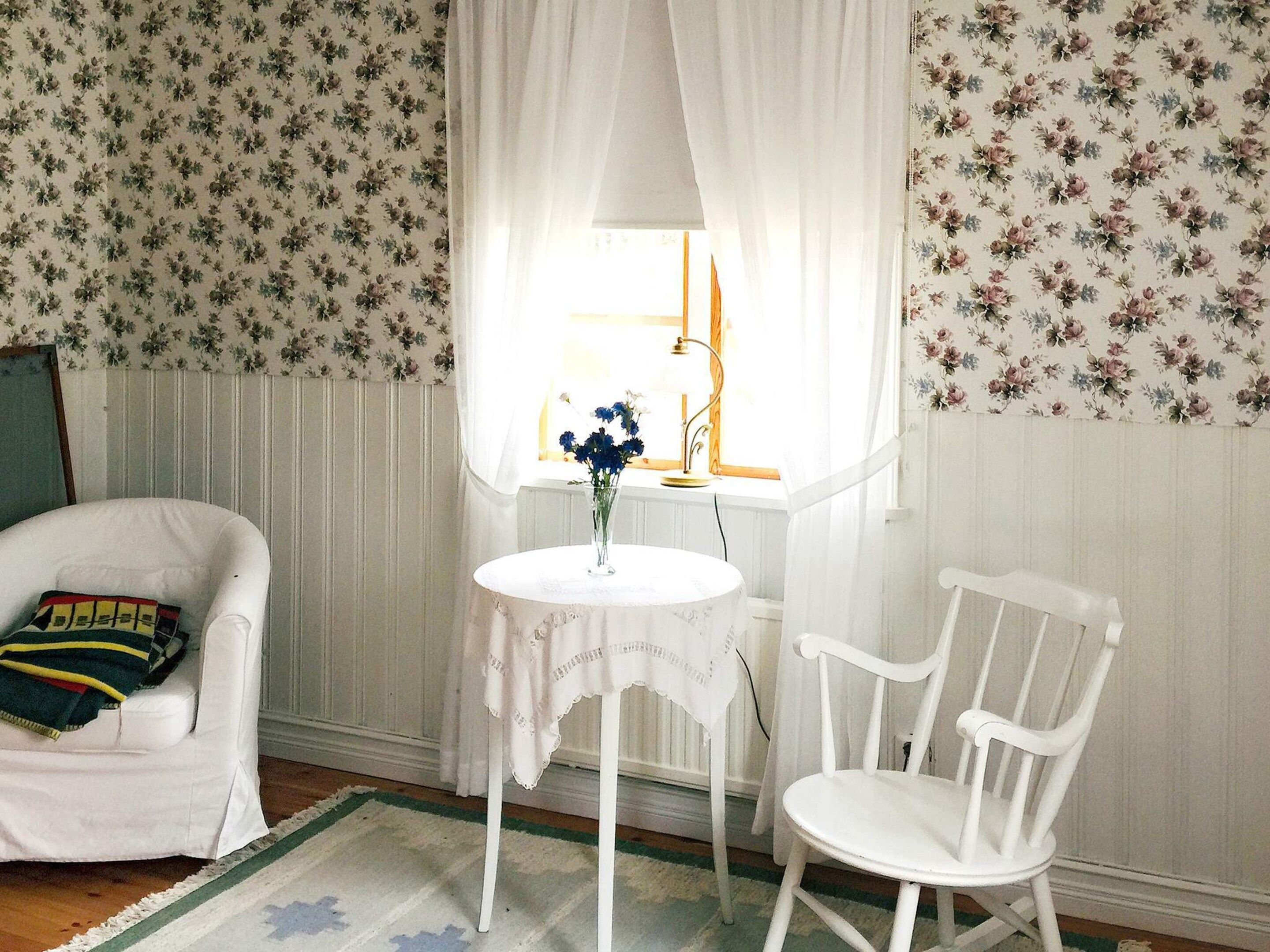 A room with floral wallpaper, a white chair, a small round table with a vase of flowers, and a window with sheer curtains.
