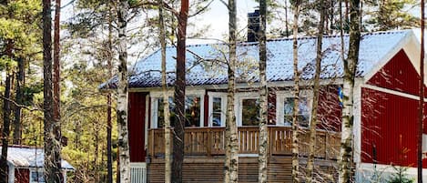 A cabin with a wooden deck, surrounded by tall trees and covered in snow.