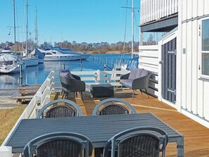 A marina with boats docked, a wooden deck with outdoor furniture, and a building with large windows. - 4 Star Holiday Home in Ebeltoft (Ebeltoft)