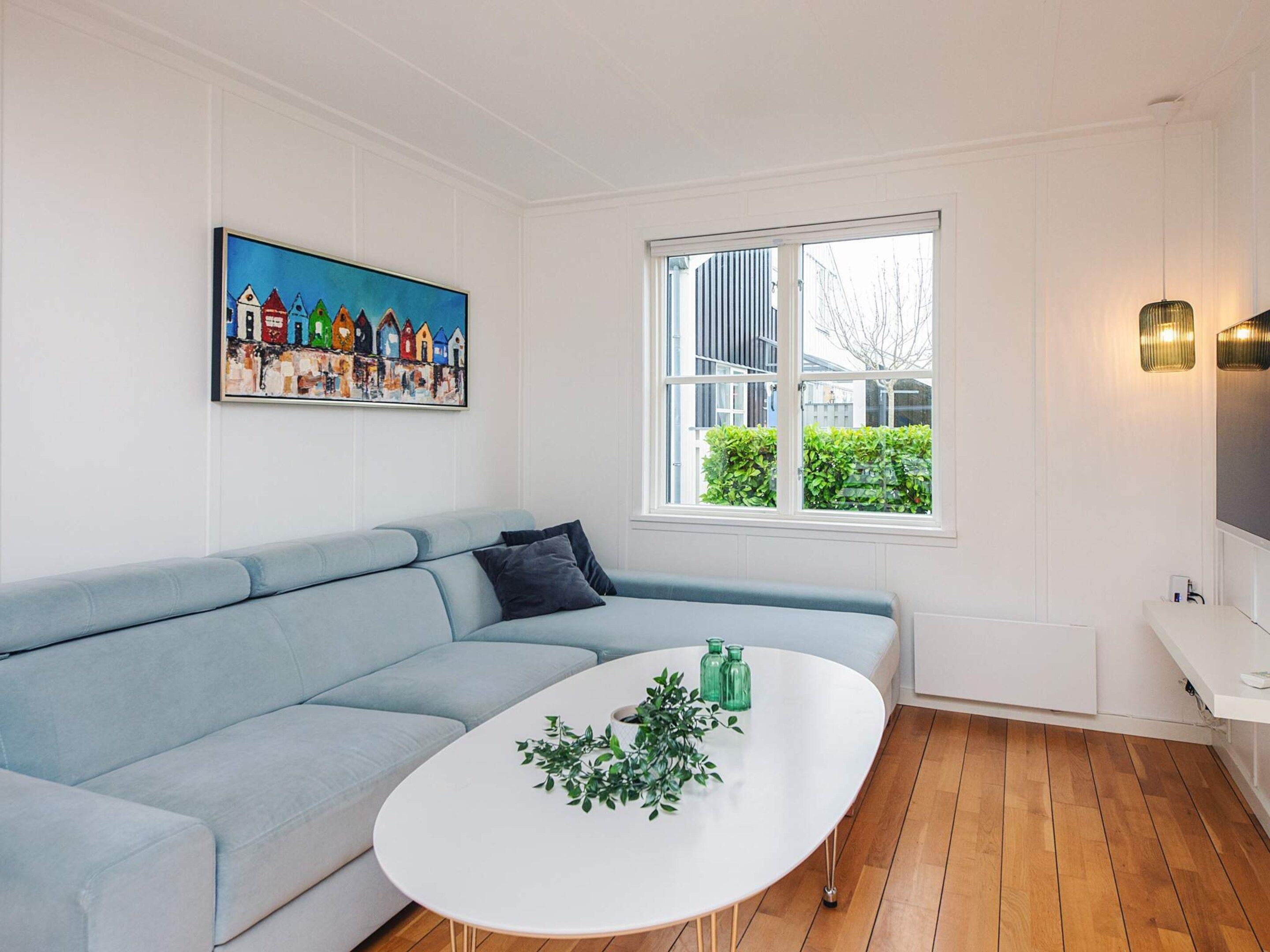 Un salón moderno con un sofá azul claro, una mesa de centro blanca con plantas verdes, un cuadro colorido en la pared y una ventana con vistas al verdor.