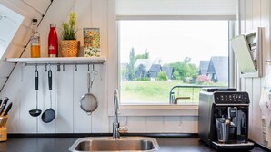 Una cocina con un lavamanos de acero inoxidable, utensilios colgantes, una cafetera y una ventana con vista a casas.