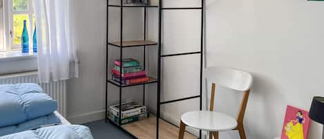 Un dormitorio con una cama, una silla blanca, un escritorio, una estantería y una ventana con cortinas.