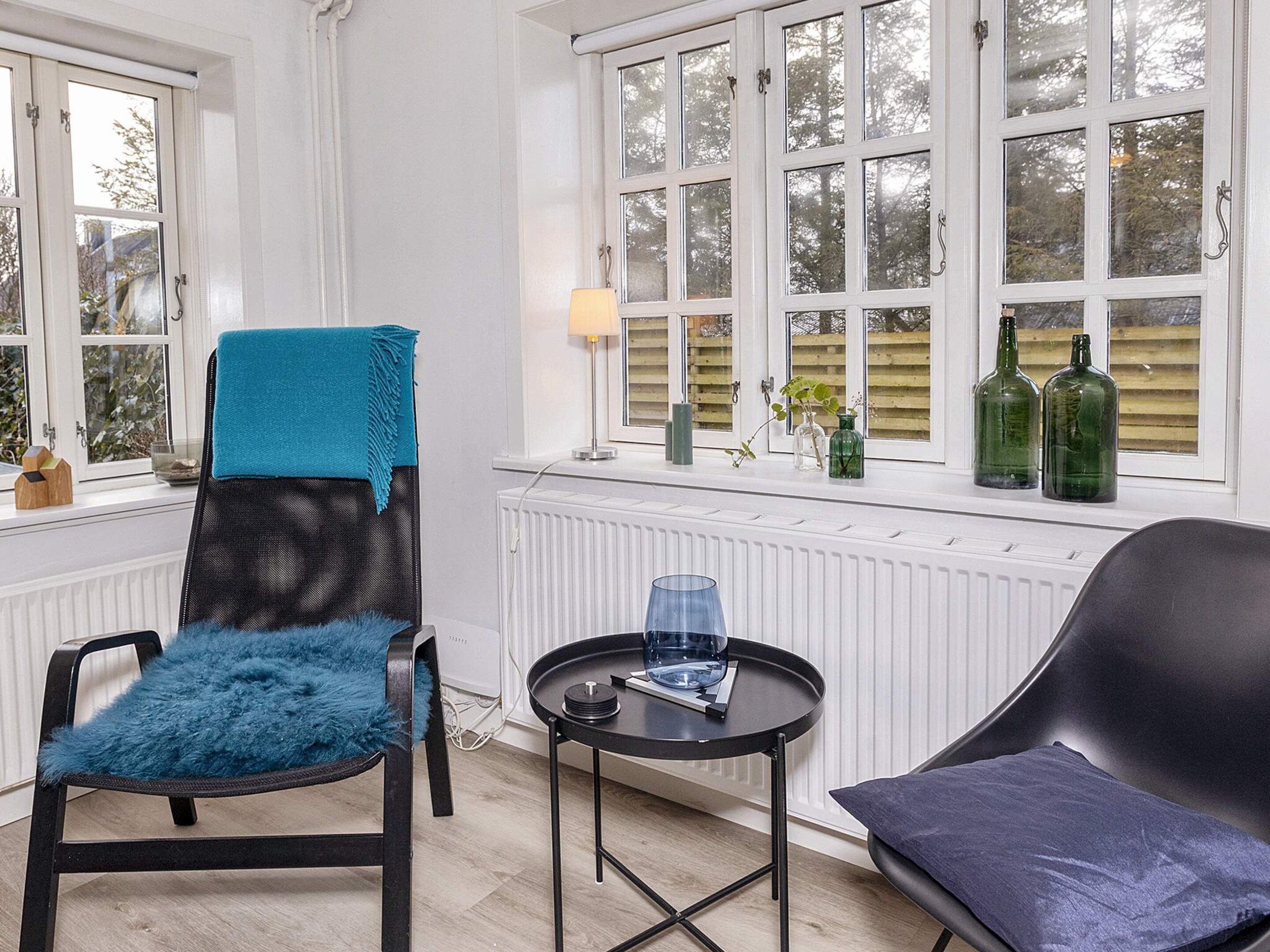 A room with a chair with a blue cover, a black chair with a dark cushion, a small round table with a glass, and a window with bottles on the sill.