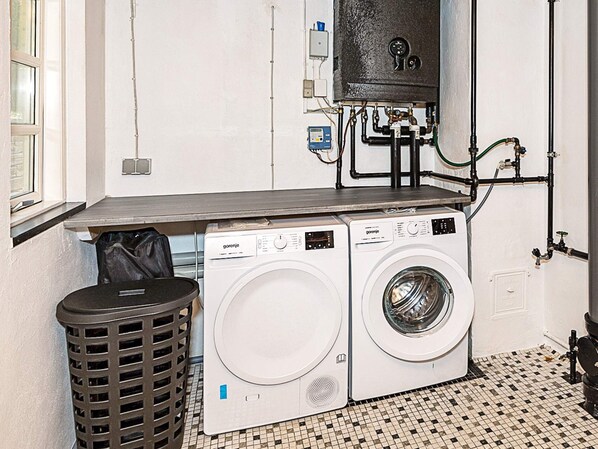 Un lavadero con dos lavadoras blancas, un bote de basura negro y un calentador de agua en la pared.