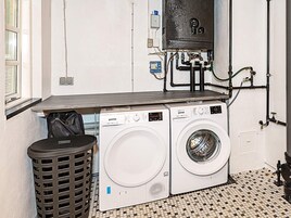 Un lavadero con dos lavadoras blancas, un cubo de basura negro y un calentador de agua en la pared.