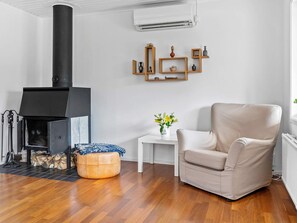 A living room with a wood-burning stove, a beige armchair, a small white table with a vase of flowers, and wooden shelves with decorative items. - 4 Star Holiday Home in Nosund (Nosund)