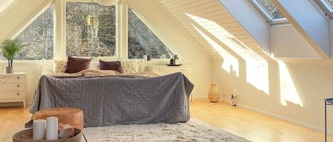 A bedroom with a sloped ceiling, large windows, a bed, bedside tables, and a bedside lamp.