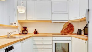 Cozinha compacta com armários brancos, bancada de madeira, forno embutido, cafeteira e rádio.