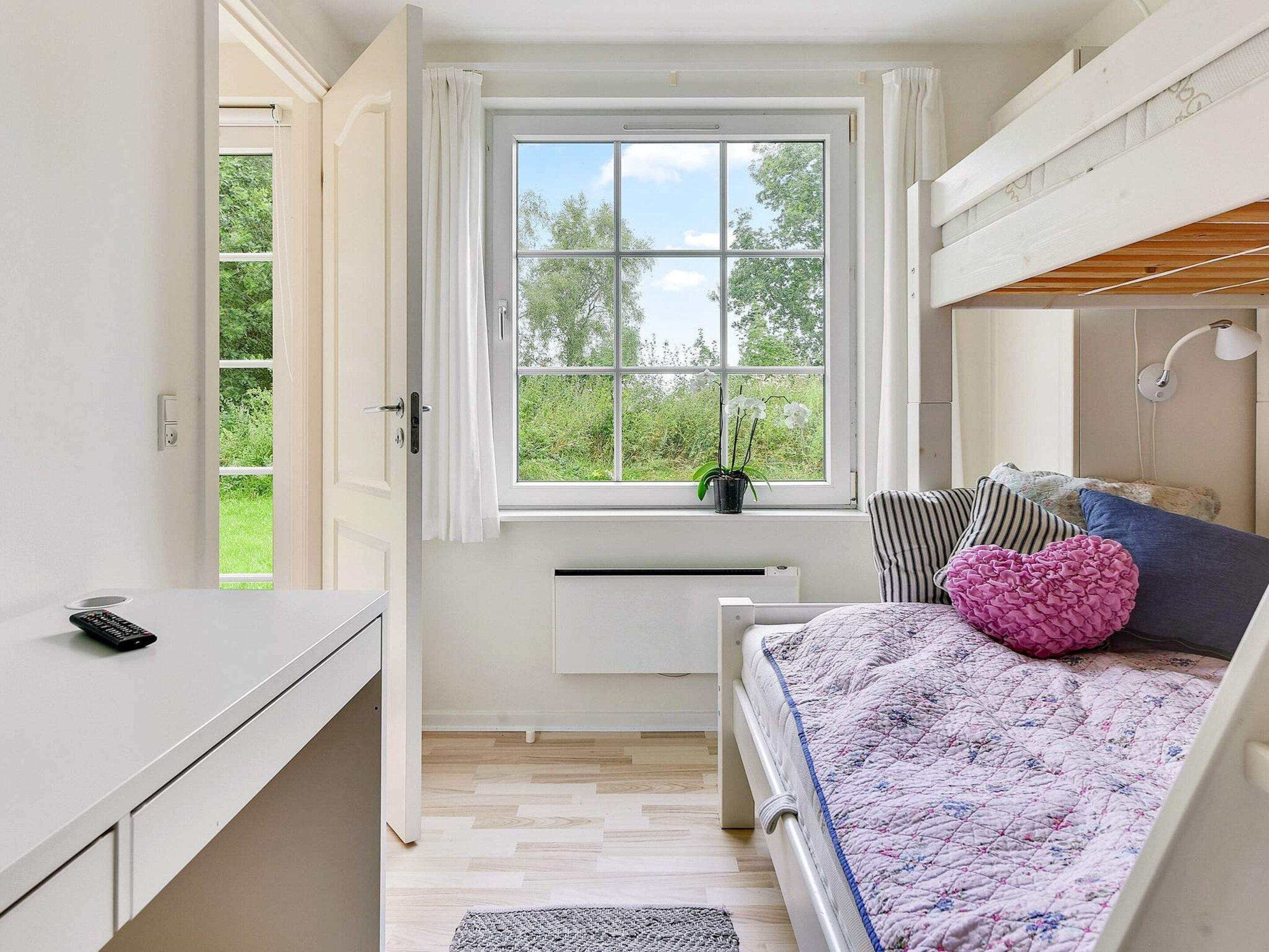 A bedroom with a bed, a desk, a window with a view of greenery, and a radiator.