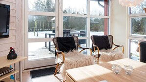 A room with a large sliding glass door leading to an outdoor area, wicker chairs with fluffy cushions, a wooden table, and a television mounted on the wall.
