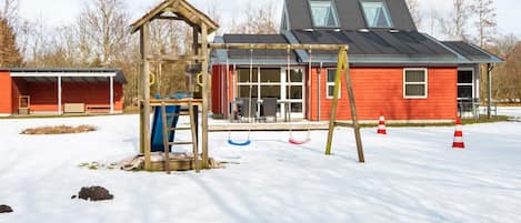 A playground with swings and a climbing structure, a red building with a covered area, and a snow-covered ground.