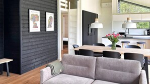 Un espacio habitable moderno con un sofá gris, una mesa de comedor de madera y sillas negras. Hay dos obras de arte enmarcadas en la pared, un banco de madera y una zona de cocina con una lámpara colgante.