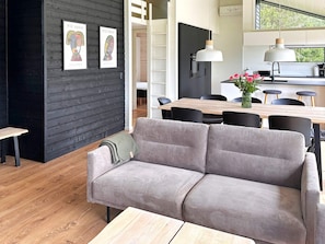 A modern living space with a grey sofa, wooden dining table, and black chairs. There are two framed artworks on the wall, a wooden bench, and a kitchen area with a hanging light fixture. - 7 Person Holiday Home in Jerup-by Traum (Jerup)