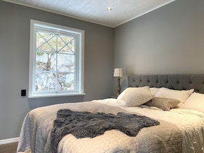 A bedroom with a large window, a bed with multiple pillows, a bedside lamp, and a dark throw blanket.