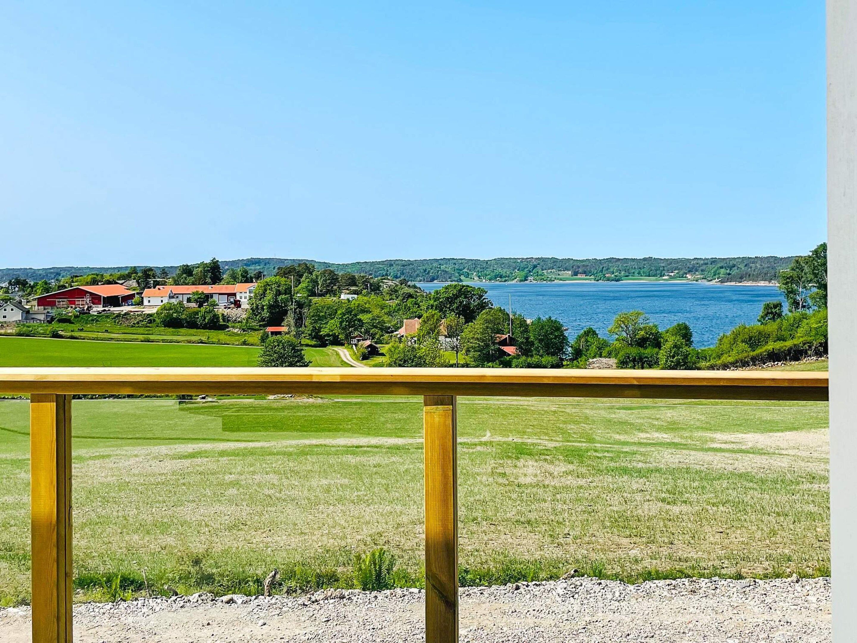 A view from a balcony with a wooden railing, overlooking a body of water, green fields, and a few buildings in the distance.