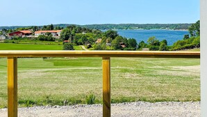 Vista desde un balcón con barandilla de madera, sobre un cuerpo de agua, campos verdes y algunos edificios a lo lejos.