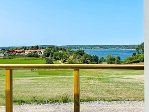 A view from a balcony with a wooden railing, overlooking a body of water, green fields, and a few buildings in the distance. - 5 Star Holiday Home in Uddevalla (Uddevalla)