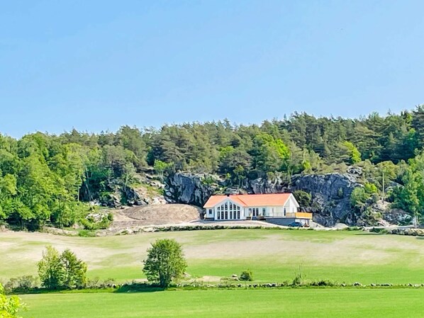 Uma casa com telhado vermelho, rodeada por uma paisagem verde e uma colina arborizada.