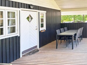A covered outdoor area with a table and chairs, a door with a window, and a dark wooden wall.