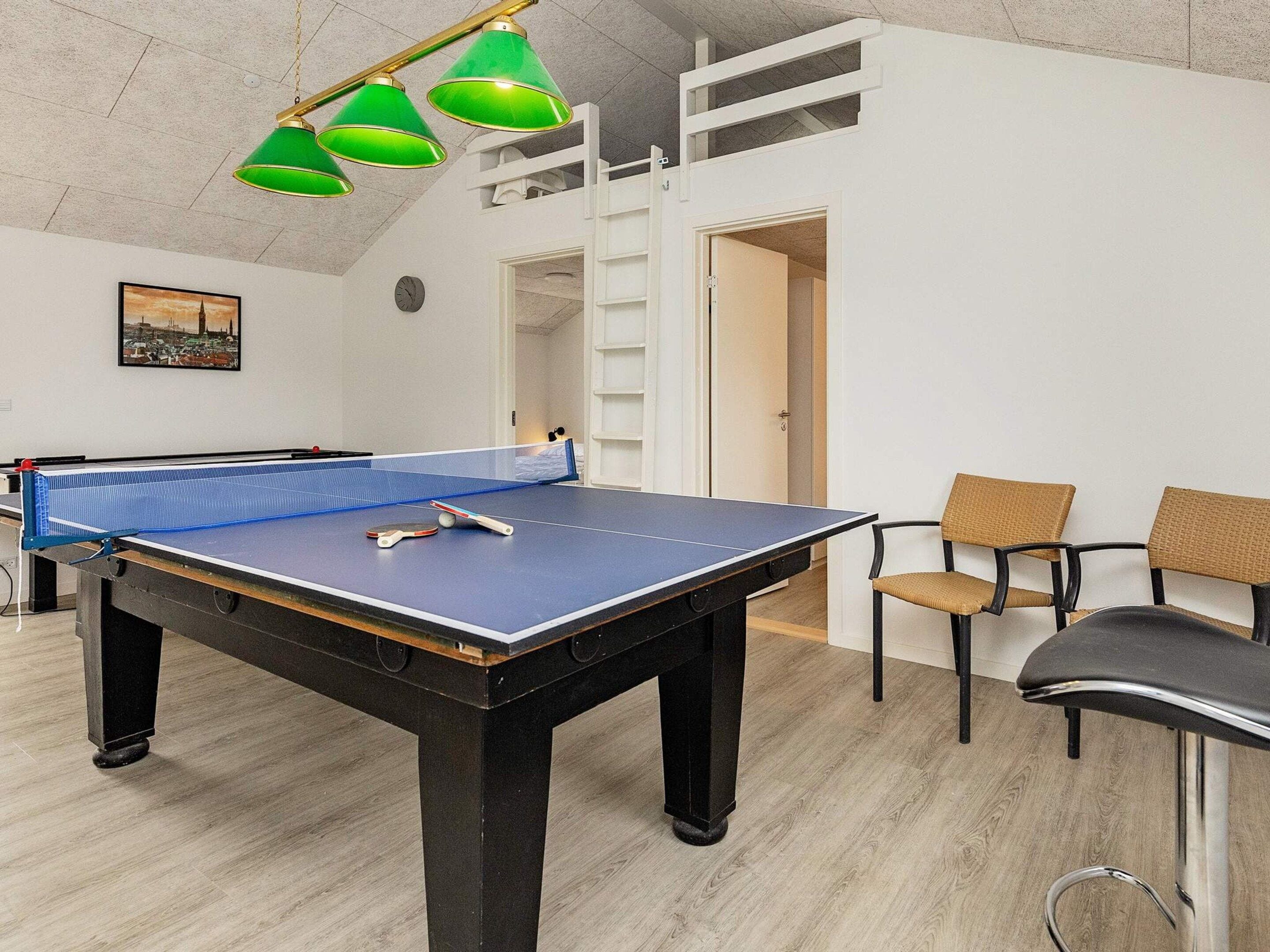 Une table de ping-pong avec un filet bleu, une horloge au mur et deux chaises en osier.