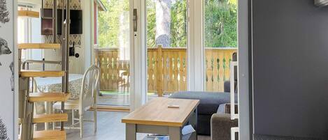 A room with a wooden table, chairs, a sofa, and a TV. There is a sliding glass door leading to an outdoor area with a wooden deck.