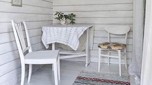A small, enclosed porch area with white wooden walls and a white table with a patterned tablecloth. There are two white chairs, one with a cushion and one without. A potted plant is on the table, and a rug is on the floor. - 4 Person Holiday Home in Axvall-by Traum (Axvall)