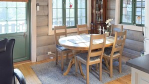 Ein Essbereich mit einem runden Holztisch, sechs Stühlen, einer Kommode und zwei Fenstern mit grünen Rahmen.