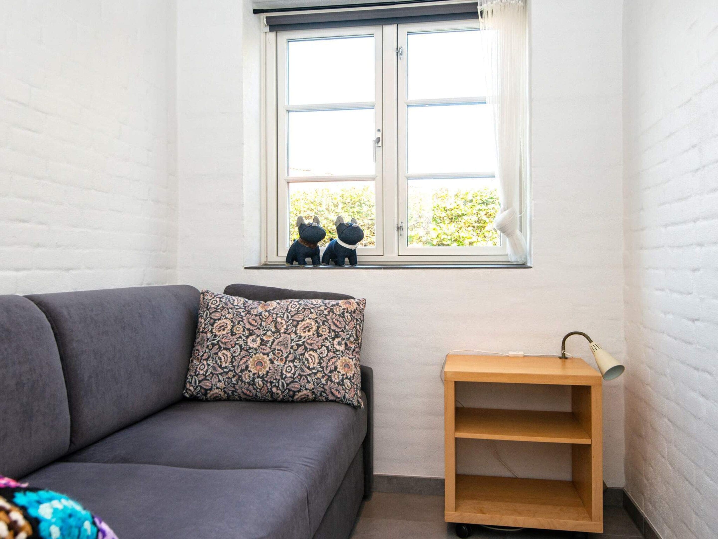 A room with a grey sofa, a patterned pillow, a small wooden shelf, and a window with two decorative dog figurines.