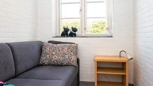 Habitació amb un sofà gris, un coixí estampat, una petita estant de fusta i una finestra amb dues figures decoratives de gos.
