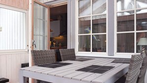 An outdoor dining area with a table and chairs, a kitchen visible through an open door, and large windows. - 3 Person Holiday Home in Munkedal (Munkedal)