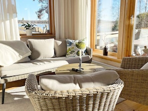 A cozy living room with a sofa, a wicker chair, a coffee table, and a view of a lake through large windows. - 4 Person Holiday Home in Hammaro (Hammaro)