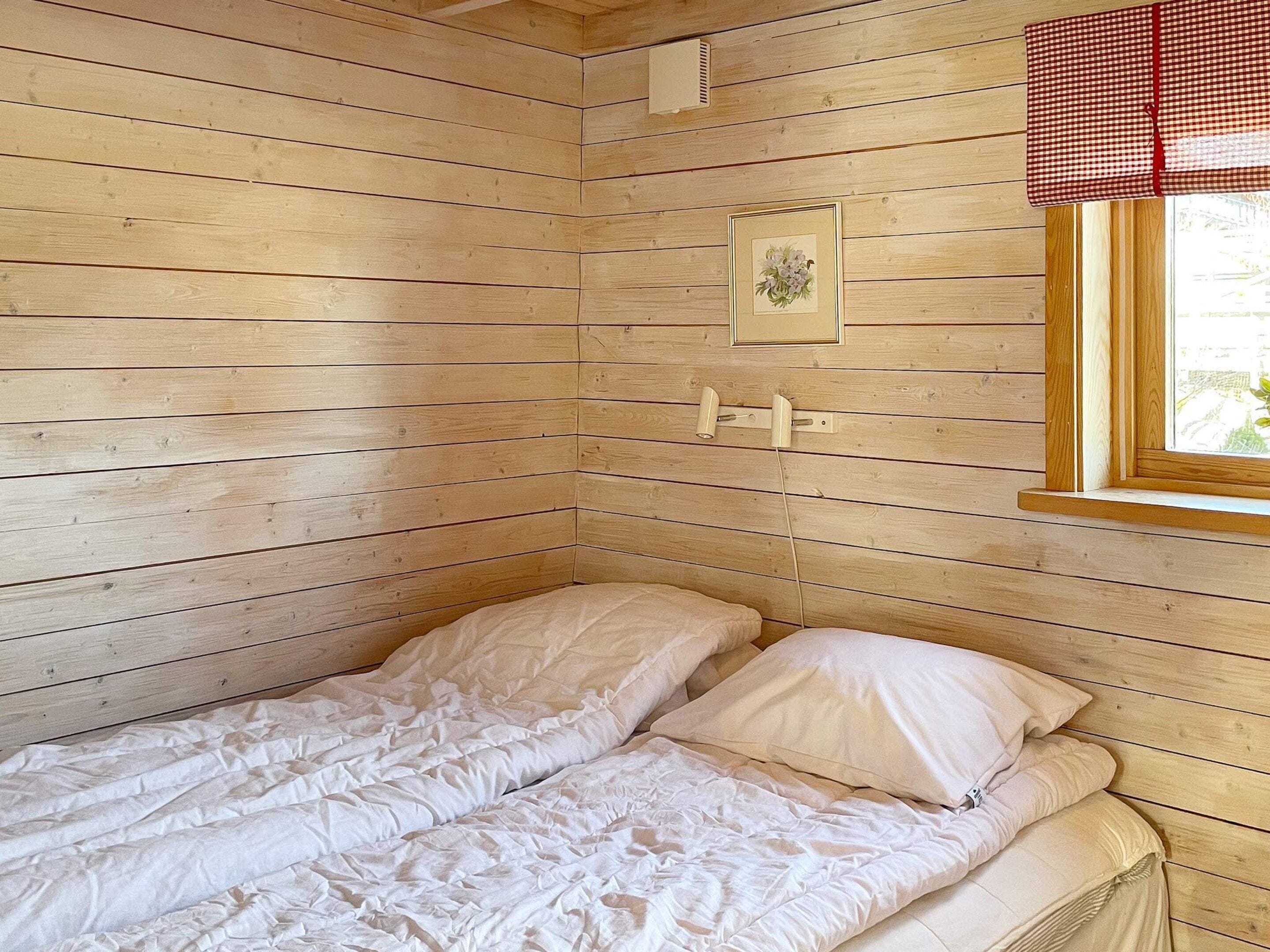 A single bed with white bedding in a room with wooden walls and a window with a checkered curtain.