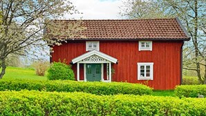 Uma casa de madeira vermelha com porta verde e detalhes brancos, cercada por sebes bem cuidadas.