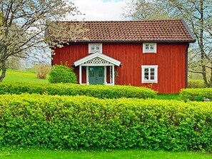 Une maison en bois rouge, avec une porte verte et des boiseries blanches, entourée de haies bien entretenues.