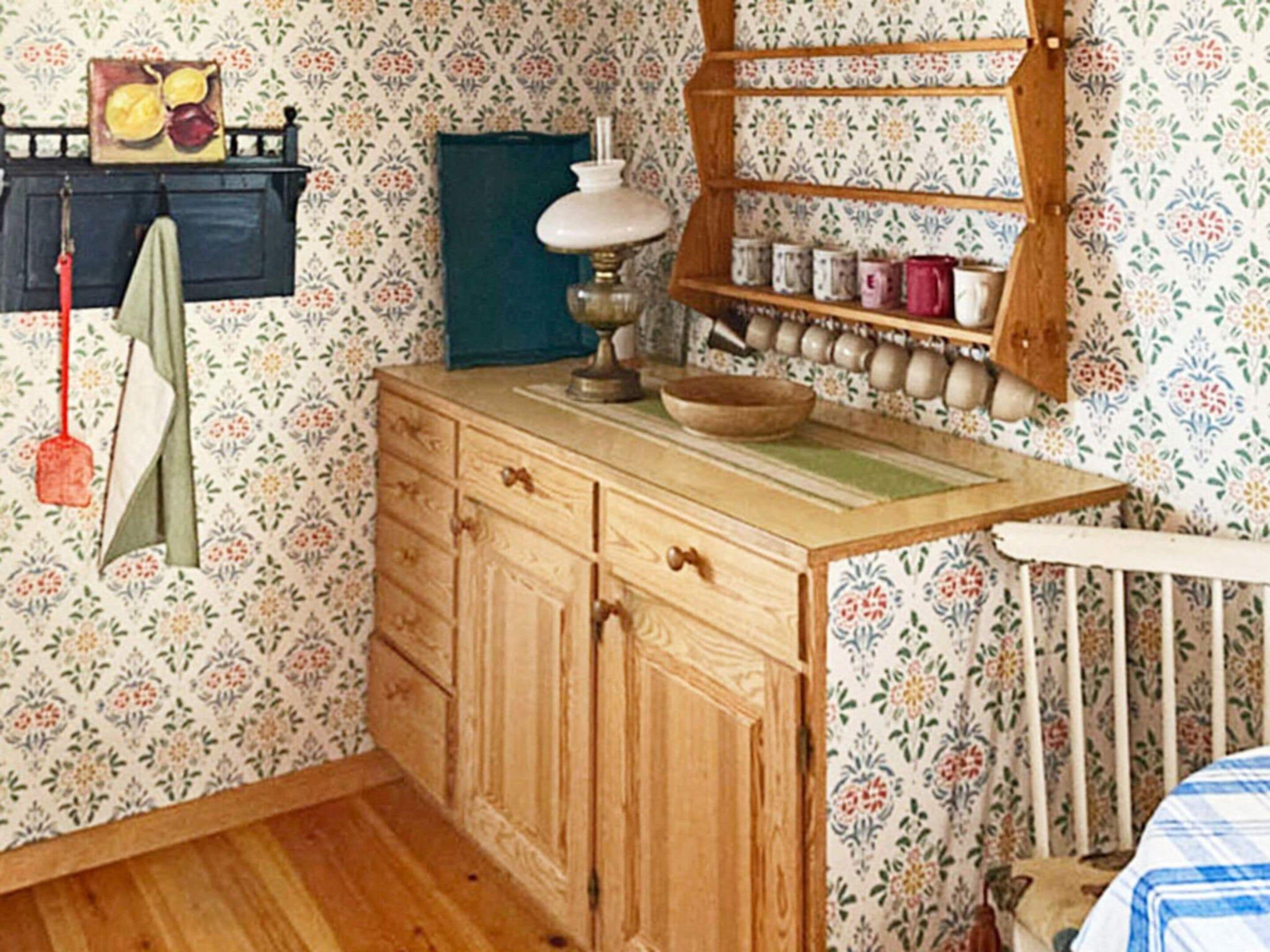 A room with a wooden dresser, a shelf with jars, a hanging towel, and a wall with floral wallpaper.