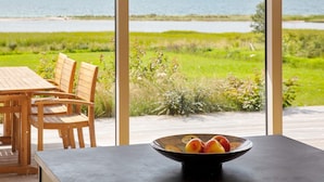 A dining area with a table and chairs, a bowl of fruit, and a view of a body of water and greenery. - 10 Person Holiday Home in Assens (Assens)