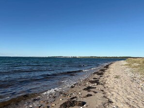 A beach with clear water, a sandy shore, and a distant shoreline with buildings. - 10 Person Holiday Home in Assens (Assens)
