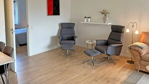 A modern living room with two grey armchairs, a small round table, a beige sofa, and a floor lamp. A white shelf with decorative items is mounted on the wall. - Seaside Bliss in Sandkas - By Traum Ferienwohnungen (Allinge)