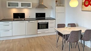 A modern kitchen with white cabinetry, a built-in oven, a microwave, and a dining area with a table and chairs. - Seaside Bliss in Sandkas - By Traum Ferienwohnungen (Allinge)
