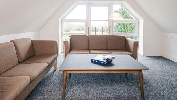 Ein Zimmer mit hohen Decken, einem Holztisch, einem braunen Sofa und großen Fenstern mit Blick ins Grüne.
