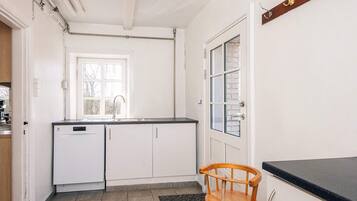 Una cocina con armarios blancos, lavavajillas, una ventana y una silla de madera.