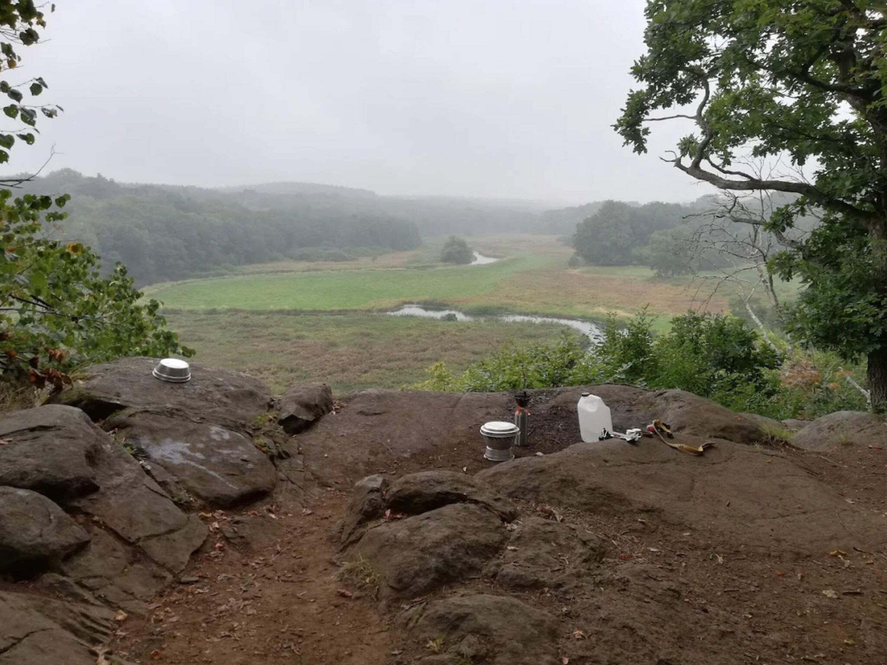 A rocky outcrop with camping gear, including a pot, a water container, and a plate, overlooking a valley with a river and distant hills.