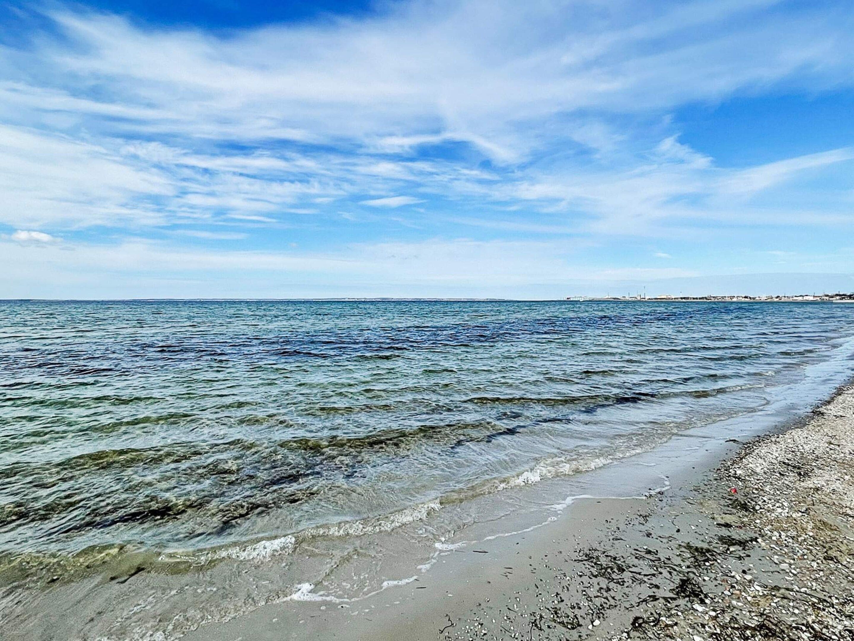 A beach with clear water, a pebbly shore, and a distant city skyline.